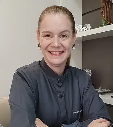 Dra. Cristiane Mariani sorrindo, de jaleco escuro, em uma das salas da clínica